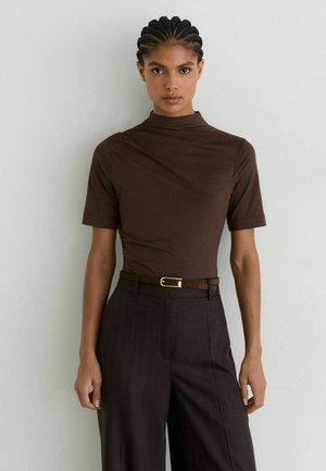 Jonge vrouw met gevlochten haar, draagt een bruin kort mouwtje met opstaande kraag en een donker broek met riem, staand tegen een lichte muur.
