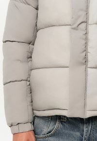 Grå polstret jakke med en glatt tekstur og kviltet design, med ribbestrikket nederkant og mansjetter. Kombinert med blå denimjeans.