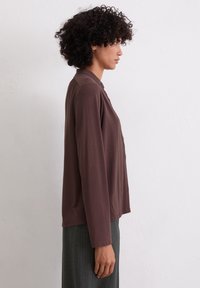 Blouse marron à manches longues avec un col et des boutons, dotée d'une coupe décontractée et d'une texture douce, portée avec un pantalon rayé gris.
