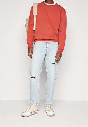 Person iført en rød sweatshirt, lyse revnede jeans, beige høje sneakers og en beige rygsæk på den ene skulder, stående mod en hvid baggrund.