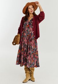 Kwiatowa maxi sukienka na ciemnym tle z różowymi i pomarańczowymi kwiatami, w zestawieniu z bordowym kardiganem i szerokim, jasnobrązowym kapeluszem.