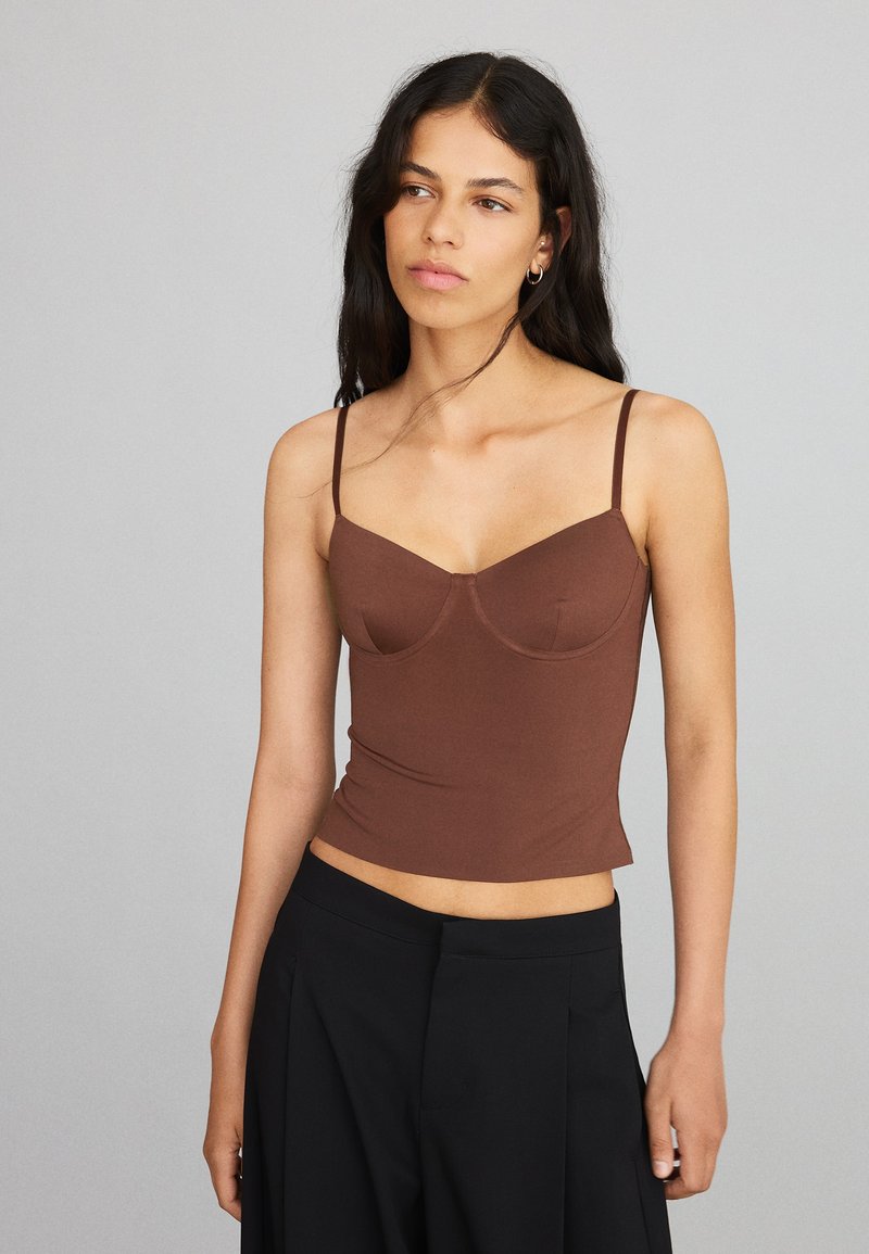 Femme aux longs cheveux foncés portant un débardeur marron ajusté et un pantalon noir ample, sur un fond uni gris clair.