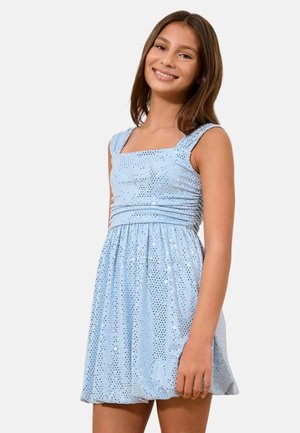 Naeratav tüdruk, kes kannab varrukateta helesinist sädelevat kleiti, millel on keha rõhutatult istuv ja lehvitav seelik, seisab ühevärvilise tausta ees.