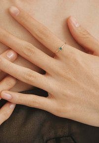 Anillo delgado de oro rosa que presenta una pequeña gema verde en el centro, utilizado en una mano con uñas naturales sobre un fondo neutro.