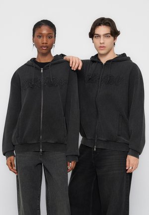 Dwie młode osoby ubrane w pasujące czarne bluzy z kapturem na zamek i ciemne dżinsy, stojące obok siebie na jednolitym jasnym tle.