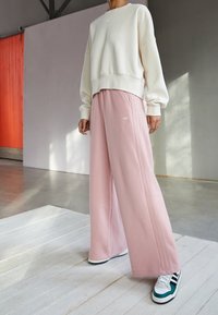 Sudadera color crema con un dobladillo acanalado, combinada con pantalones deportivos sueltos rosas que tienen rayas laterales y un pequeño logotipo. El calzado incluye zapatillas con detalles verdes.