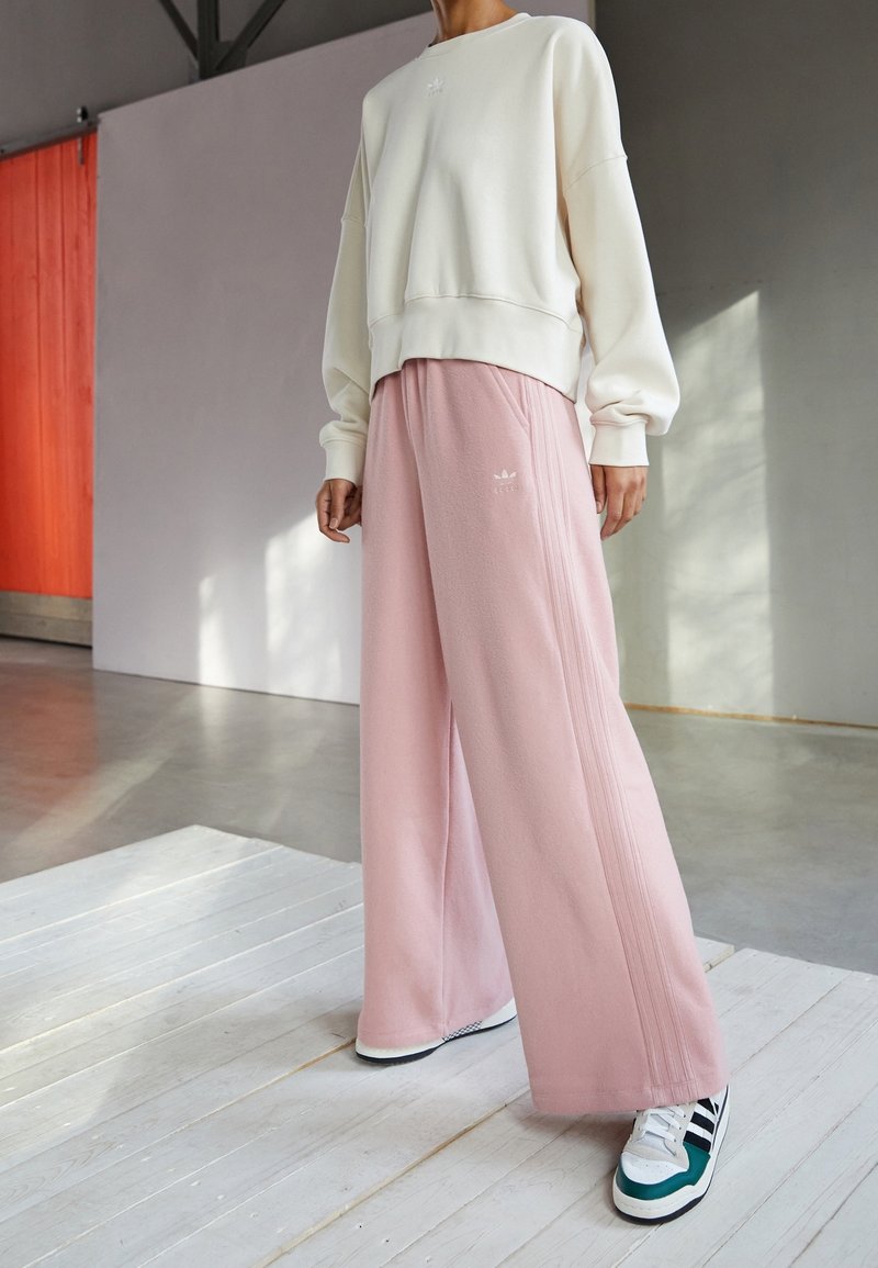 Sudadera color crema con un dobladillo acanalado, combinada con pantalones deportivos sueltos rosas que tienen rayas laterales y un pequeño logotipo. El calzado incluye zapatillas con detalles verdes.