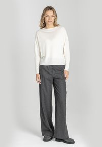 Kobieta stoi w białej bluzce z długim rękawem, szarych szerokich spodniach i ciemnych butach na tle jednolitego jasnoszarego tła.