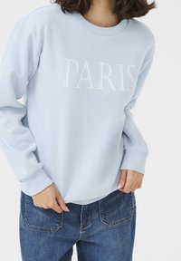 Felpa azzurra con la scritta "PARIGI" in bianco. Il design include polsini a coste e un collo tondo, abbinata a jeans blu.