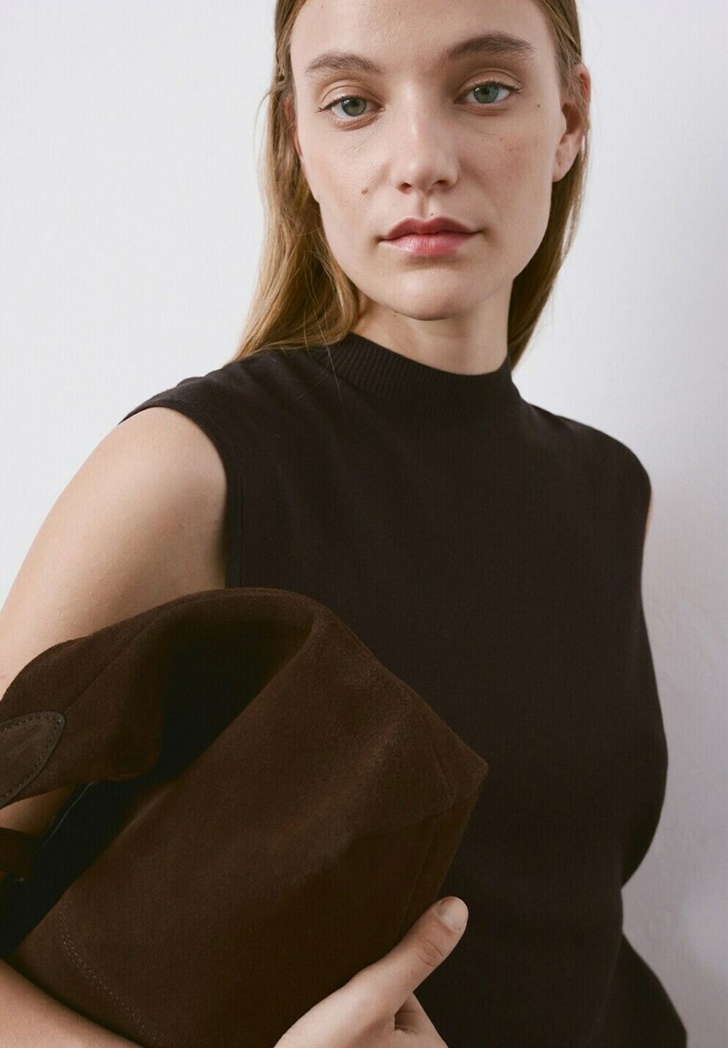 Jonge vrouw met een lichte huid en groene ogen, gekleed in een mouwloos zwart topje, met een donkerbruine suède jas over haar arm.