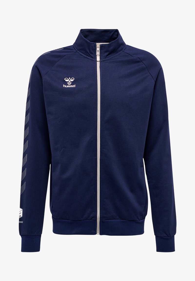 Veste zippée navy en tissu lisse. Dotée d'un col montant, d'une fermeture éclair blanche et de rayures en chevron blanches sur les manches. Logo Hummel sur la poitrine.