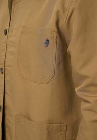 Chaqueta de camisa de algodón color tostado con cierre de botones, bolsillo en el pecho y logo bordado en azul, con una textura suave y un diseño resistente.
