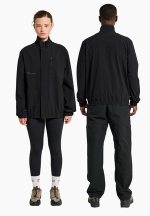 Femme de face et homme de dos portant des vestes noires assorties, pantalons noirs, chaussettes claires et chaussures de randonnée sur fond blanc.