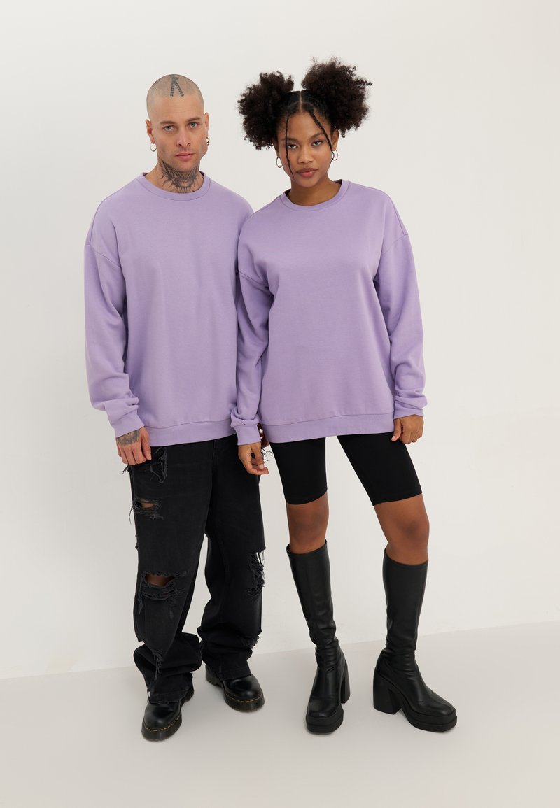 Felpe in lavanda a vestibilità rilassata, indossate da due modelli. Il modello maschile abbina un paio di jeans neri strappati; la femminile con leggings neri e stivali alti fino al ginocchio.
