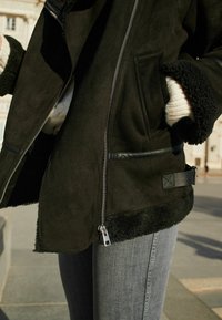 Zwarte suède jas met pluche zwart bontaccenten, voorzien van een zilveren rits, zijzakken en leren afwerkingen aan de zoom en taille.