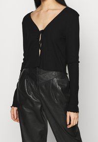 Schwarzes, geripptes Langarm-Top mit tiefem V-Ausschnitt und Bindendetail vorne, kombiniert mit hoch taillierter schwarzer Lederhose.