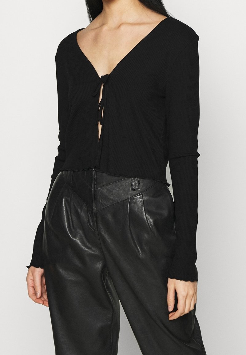 Schwarzes, geripptes Langarm-Top mit tiefem V-Ausschnitt und Bindendetail vorne, kombiniert mit hoch taillierter schwarzer Lederhose.
