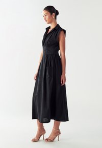 Črna brezrokavnik obleka s strukturiranim ovratnikom, empire pasom, nabranim krilom in sprednjim žepom, v kombinaciji z nude čevlji na visoki peti z naramnicami.