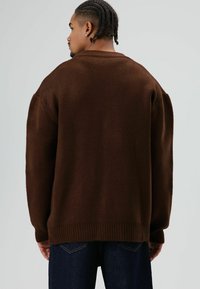 Pull en tricot marron à coupe décontractée, avec poignets et ourlet côtelés, présentant une surface texturée et des épaules tombantes. Porté avec un jean foncé.