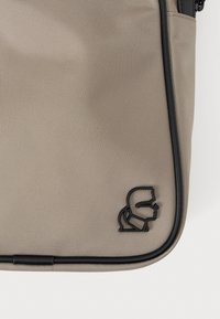 Beige nejlon táska texturált felülettel, fekete cipzárral és fekete logóval. Lekerekített élek és minimalista dizájn.