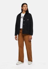 Black cotton jacket with a collared neck, button closures, side pockets, worn over a white graphic t-shirt and brown cargo pants.
