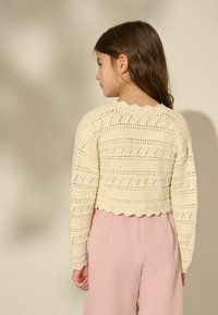 Cardigan crema a maglia con motivo a giorno e orlo scallop, indossato sopra pantaloni rosa chiaro. Presenta maniche lunghe e scollatura rotonda.