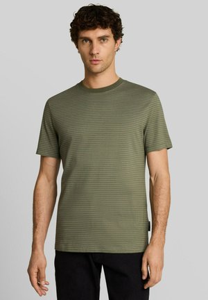 Mann mit lockigem Haar, der ein olivgrün gestreiftes kurzärmeliges Hemd und schwarze Hose trägt, steht vor einem schlichten grauen Hintergrund.