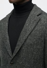 Hallhalline herringbone bleiser, millel on notšitatud krae ja üksik nupp, koos musta kõrge kaelusega särk. Omab krael väikest metallaktsenti.