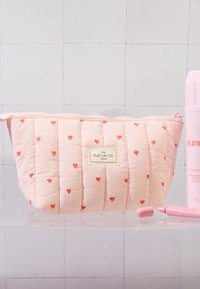 Quiltet rosa kosmetikkveske med små røde hjerter, en etikett som leser "THE FLAT LAY CO. LONDON," og glidelås.