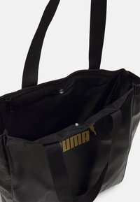 Sac fourre-tout Puma noir avec logo doré, surface texturée, poignées doubles et ouverture supérieure zippée montrant la doublure intérieure et la fermeture à bouton-pression.