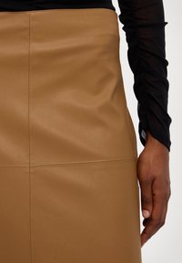 Ruskea nahkainen lyhyt hame, jossa näkyvät saumat, sileä pinta ja kapea leikkaus. Käsi on laskostuksen päällä, yhdistettynä mustaan yläosaan.