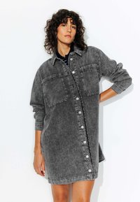 Robe chemise en denim gris avec un devant à boutons, deux poches poitrine, des manches longues et une coupe décontractée, avec une texture délavée.