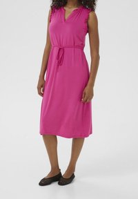 Robe sans manches rose avec des épaules volantes, décolleté en V et taille à cordon. Ourlet droit, longueur genou. Porte avec des ballerines noires texturées.