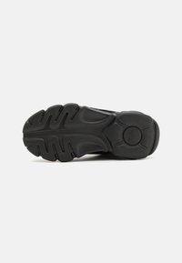 Semelle de chaussure noire avec des rainures profondes et un profil texturé, conçue pour l’adhérence et la durabilité, présentée sur un fond blanc uni.