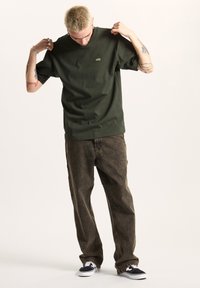 Camiseta de manga corta verde oscuro con un pequeño logo amarillo de Vans, combinada con pantalones vaqueros marrones holgados y zapatillas en blanco y negro.