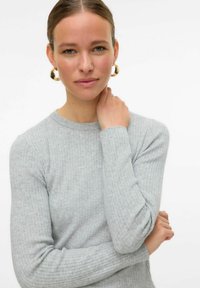 Šviesiai pilkas raštuotas megztinis su ilgomis rankovėmis, apvaliu iškirpimu ir švelnia tekstūra. Modelis su auksinėmis auskarų lankomis liečia savo kaklą.