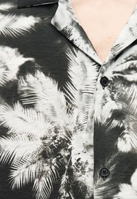 Kortermet skjorte i svart og hvitt med palmebladprint. Har krage og svarte knapper langs fronten, teksturert stoff.