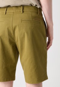 Olivenfargede shorts med en glatt tekstur, standard beltehemper og to baklommer. Har et merketagg over høyre lomme.