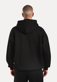 Uomo con capelli corti che indossa un semplice hoodie nero e pantaloni neri, in piedi rivolto di spalle su uno sfondo semplice.
