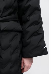 Crna puffer jakna sa quilted teksturom, koja ima pojas u struku, bočni džep i bijeli Nike logo na rukavu.
