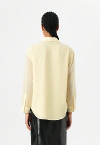 Camicia gialla a maniche lunghe realizzata in un tessuto leggero e trasparente, con colletto classico e polsini abbottonati, indossata sopra una gonna in pelle nera.