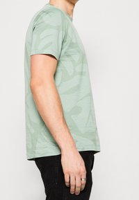 Vaaleanvihreä t-paita, jossa on teksturoitu kangas ja hienovarainen camo-kuvio; lyhyet hihat, pyöreä kaula-aukko ja rento mitoitus.