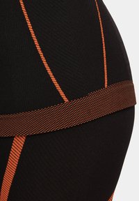 Czarne legginsy sportowe z teksturowanej tkaniny, z żywymi pomarańczowymi paskami i wzorzystym elastycznym pasem w talii. Zbliżenie na szczegóły.