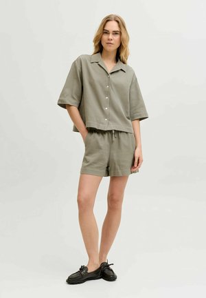 Donna in piedi che indossa una camicia verde oliva ampia con bottoni e pantaloncini abbinati con scarpe slip-on nere su uno sfondo chiaro e semplice.
