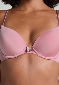 Lichtroze gewatteerde beha met dunne verstelbare bandjes, voorzien van een klein strikje en logo-detail in het midden. Glad oppervlak overal.