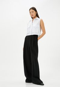 Kort wit button-up vest met twee voorkwasten, gecombineerd met hoge zwarte wijde broekspijpen, voorzien van een soepele textuur en subtiele plooien.