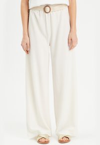 Wide-leg, textured cream pants with a braided belt featuring a circular buckle. Worn with gold-tone flat sandals.