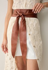 Brown leather belt with a large bow, worn over a cream, lace-textured garment. Features long, smooth ribbon ends and a structured design.