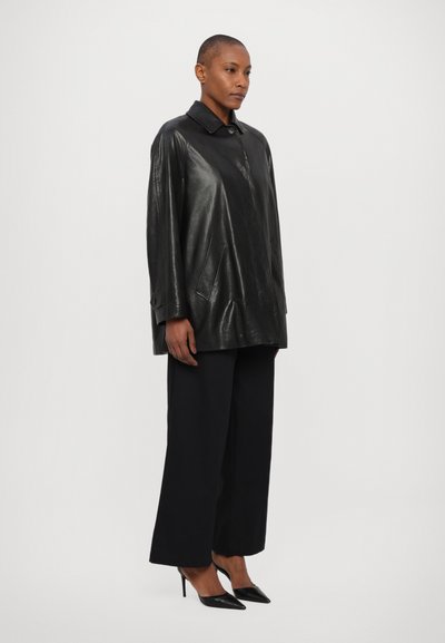 Camicia overshirt in pelle nera con colletto, bottoni frontali e tasche laterali; abbinata a pantaloni larghi neri e décolleté neri appuntiti.