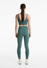 Tealfärgad sport-bh och matchande högmidjade leggings med rynkdetaljer. Modell med vita träningsskor, sedd från ryggen.